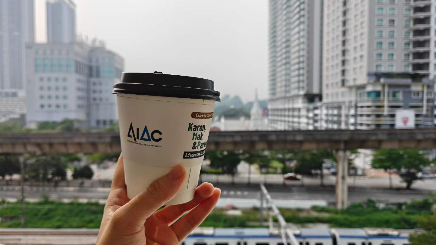A hand holding an AIAC-branded coffee cup against a city backdrop