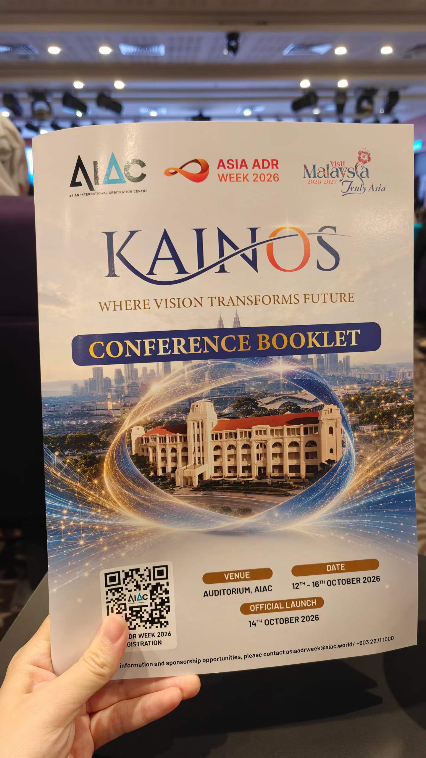 A hand holding the KAINOS conference booklet for Asia ADR Week 2026 at the AIAC Auditorium in Kuala Lumpur