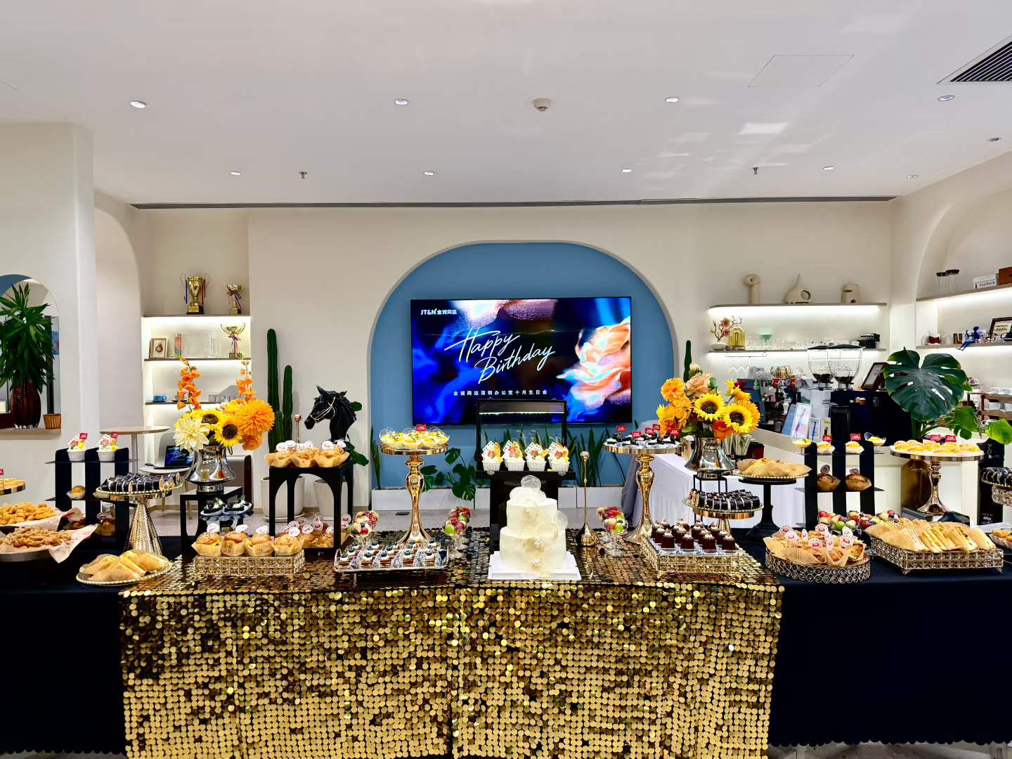 A dessert table with a white birthday cake, pastries, sunflowers, and a Happy Birthday screen at the JT&N October birthday celebration